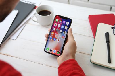Fotografie osoby sedící u stolu s šálkem kávy, drží iPhone X. Na obrazovce iPhone X je zobrazena řada ikon aplikací.