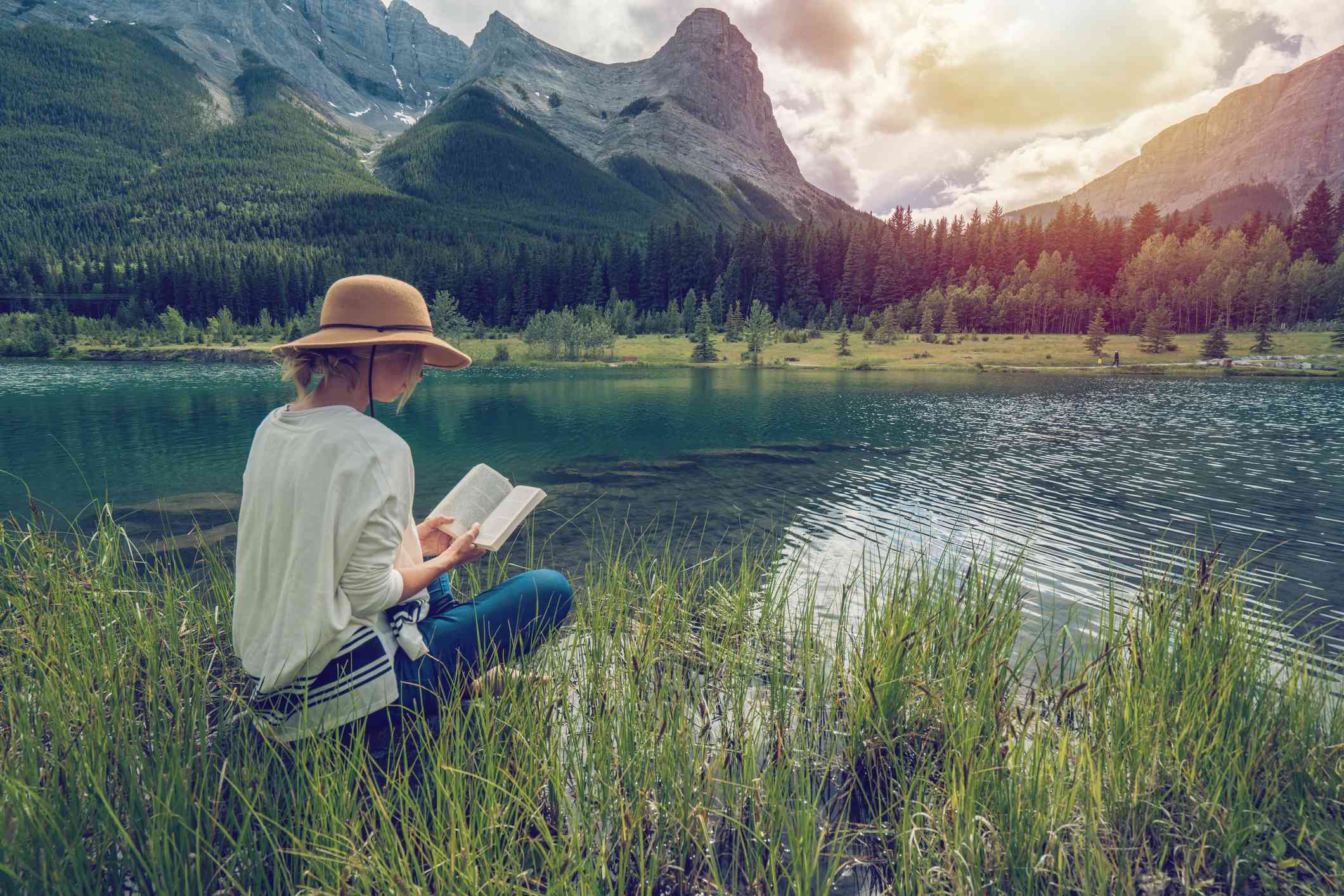Žena čte brožovaný výtisk u jezera s horami v pozadí.