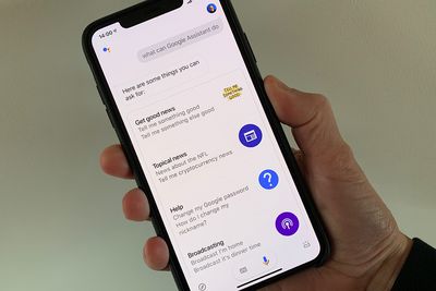 Fotografie z držení zařízení iPhone XS Max se snímkem obrazovky s akcemi Google Assistant (např. Získat dobré zprávy, Aktuální zprávy, Nápověda atd.)