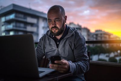 Holohlavý a vousatý muž používá notebook a smartphone při západu slunce.