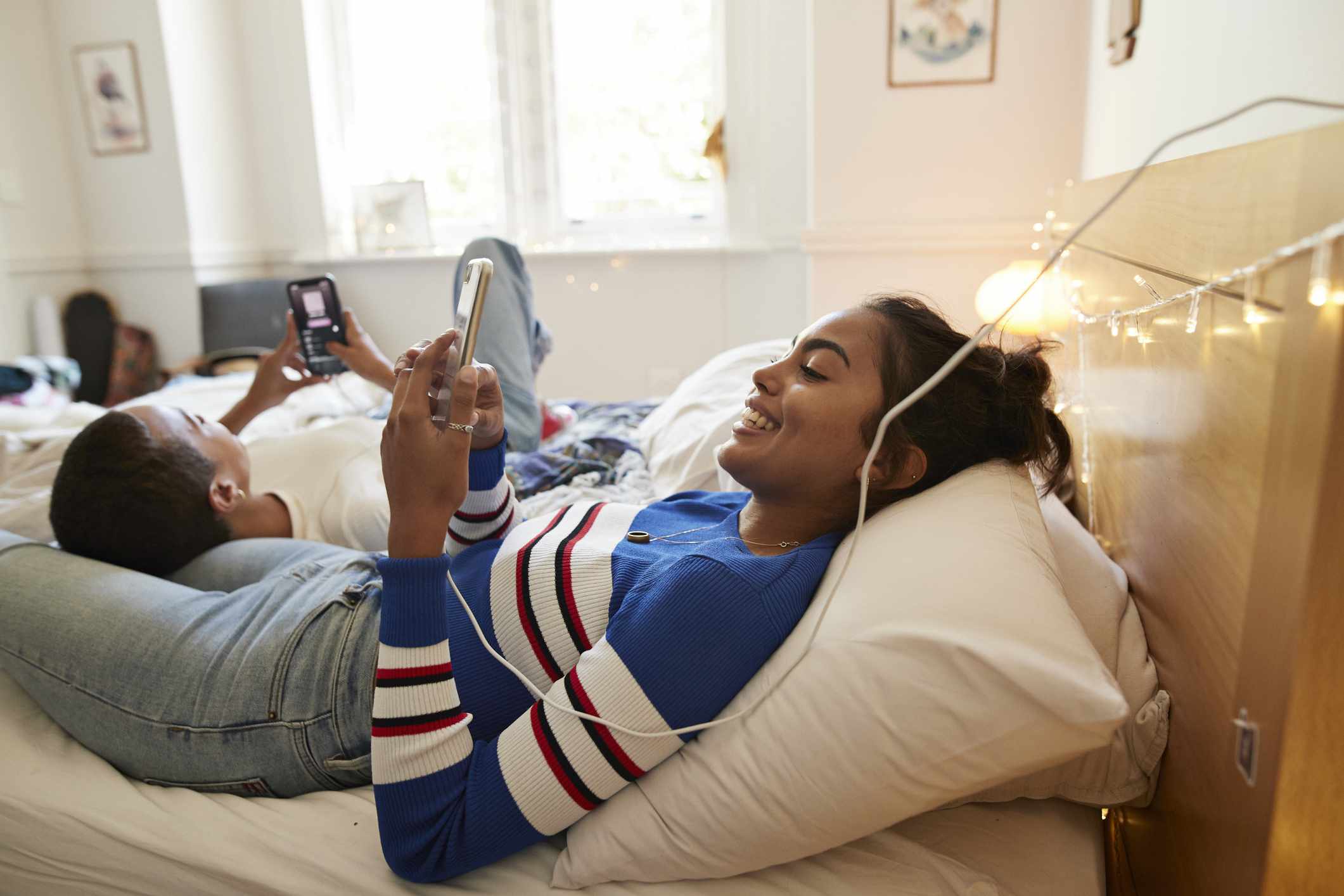 Dvě dospívající ležící na posteli pomocí svých smartphonů, zatímco jedna se nabíjí prostřednictvím připojeného kabelu.