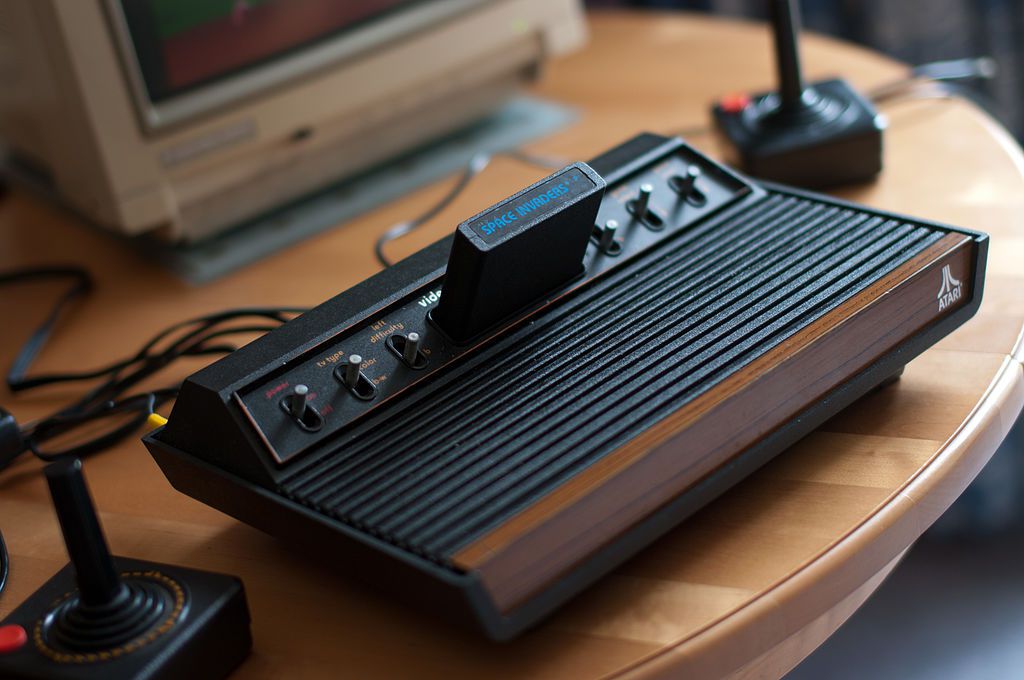Konzole Atari 2600 VCS, plné zobrazení, s joysticky a monitor Commodore 1084S částečně viditelný.  1978 model se šesti přepínači.  Hra Space Invaders je vložena.
