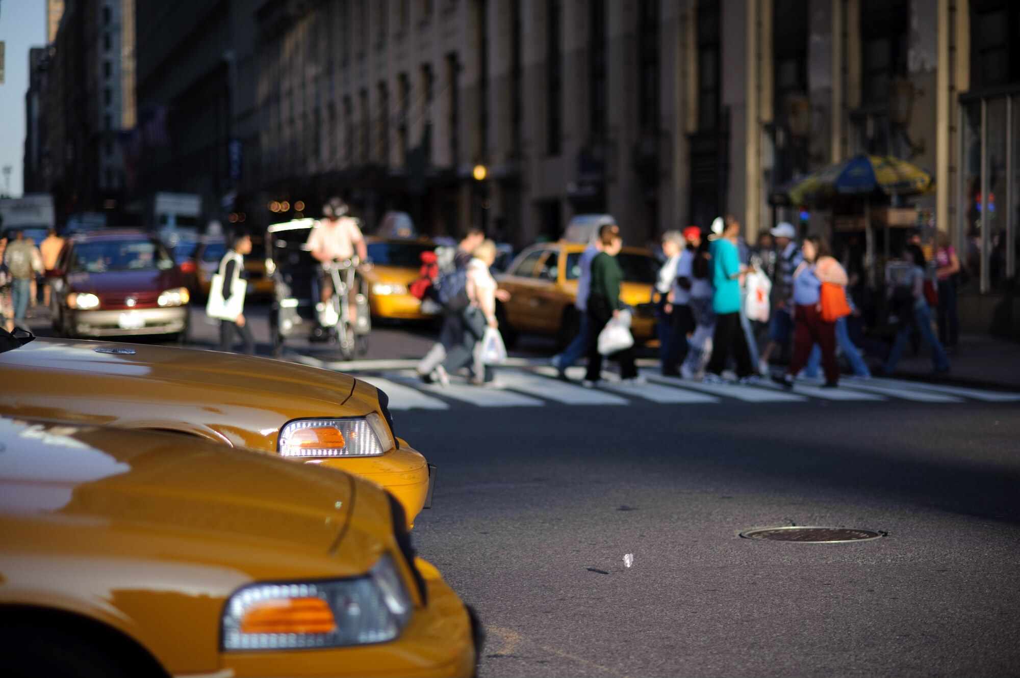 Dvojice taxíků čekajících na křižovatce, zatímco lidé v pozadí přecházejí ulici