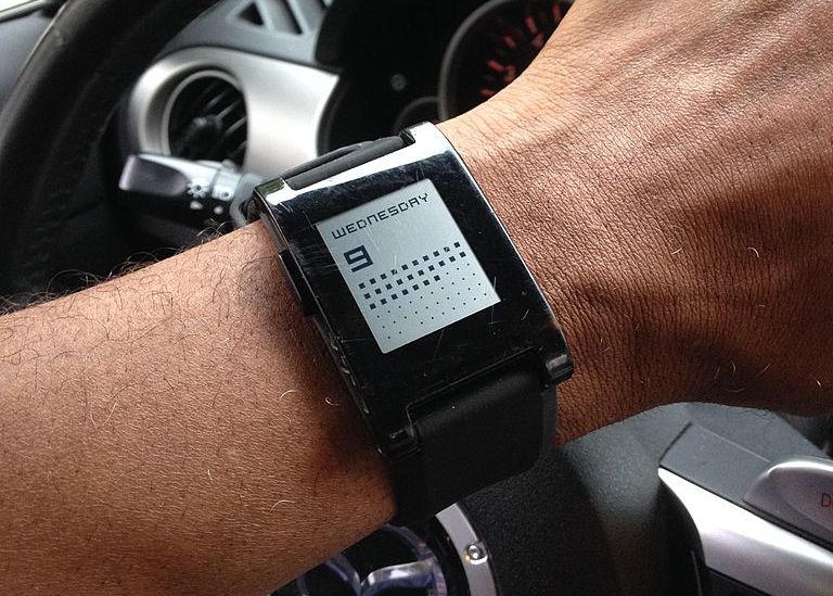 Detailní záběr na hodinky Pebble na něčí zápěstí s automobilovými displeji v pozadí. 