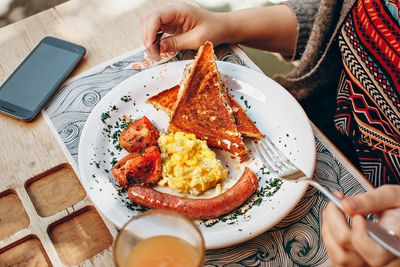 Zblízka aperson jíst snídani: toast, vejce, rajčata a klobása na talíři.  Na stole vedle talíře je vypnutý smartphone otočený lícem nahoru.