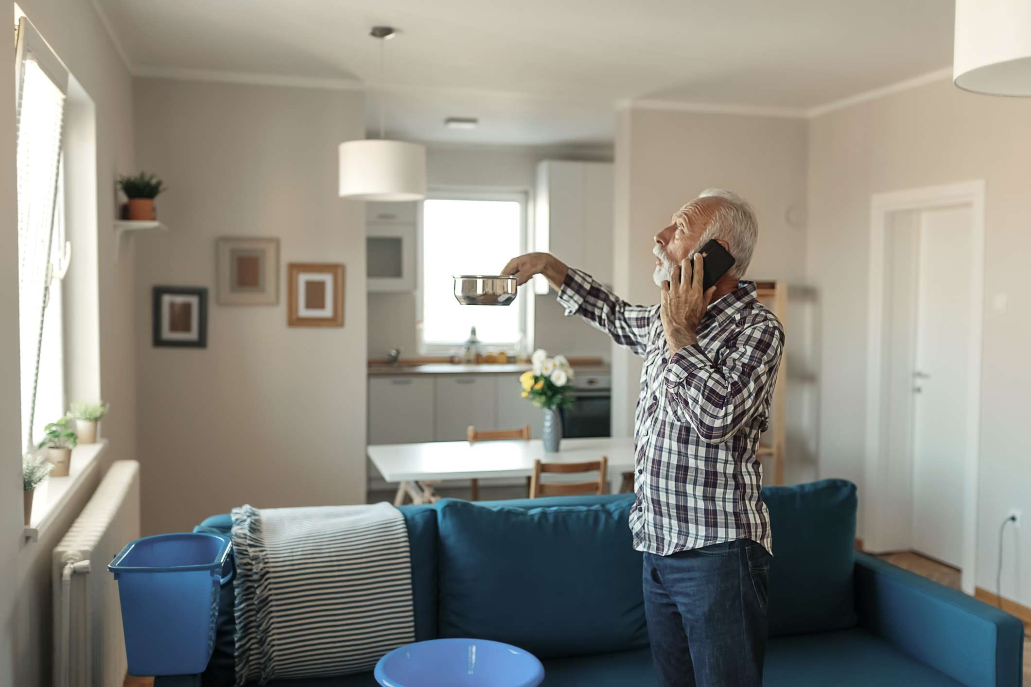 Vousatý starší muž mluví na smartphone poblíž plastové umyvadlo doma v obývacím pokoji kvůli úniku střechy