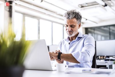 Zralý podnikatel sedí u stolu v kanceláři při pohledu na smartwatch