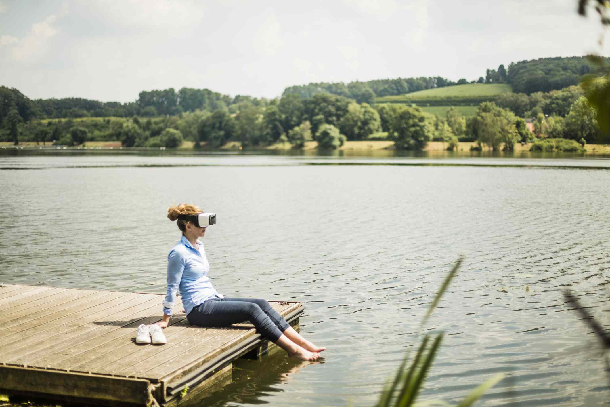 Někdo s brýlemi VR sedí na molu u jezera s nohama ve vodě. 