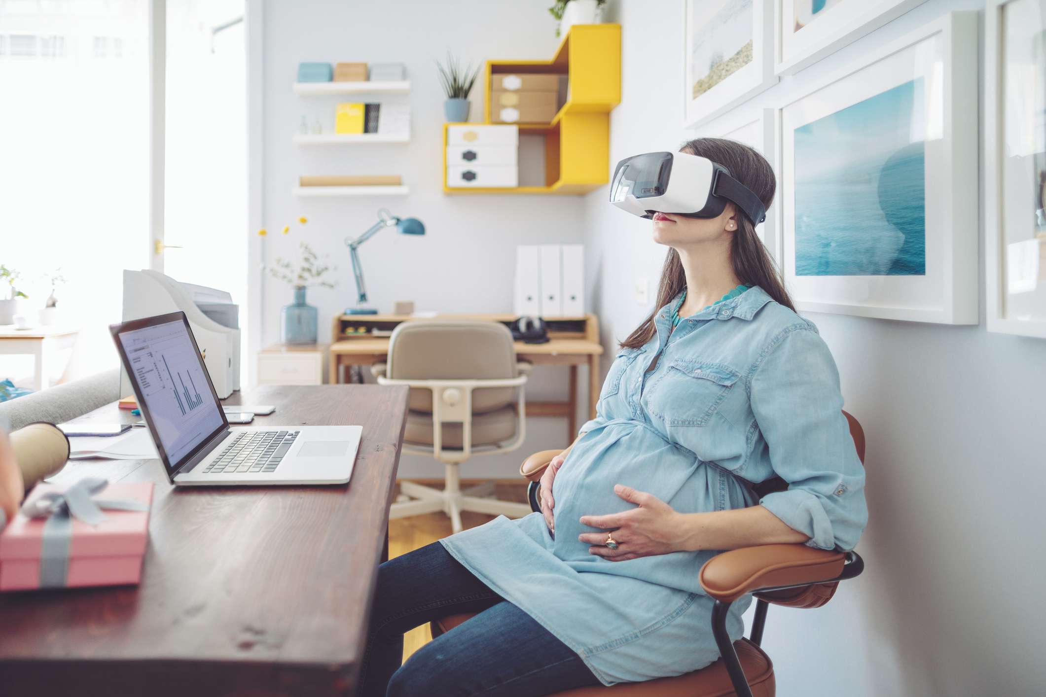 Těhotná žena nosí sluchátka VR při práci v domácí kanceláři s notebookem v okolí.