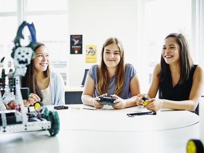 Usměvavá studentka sedí u stolu v učebně střední školy se spolužáky pomocí dálkového ovládání k ovládání robota