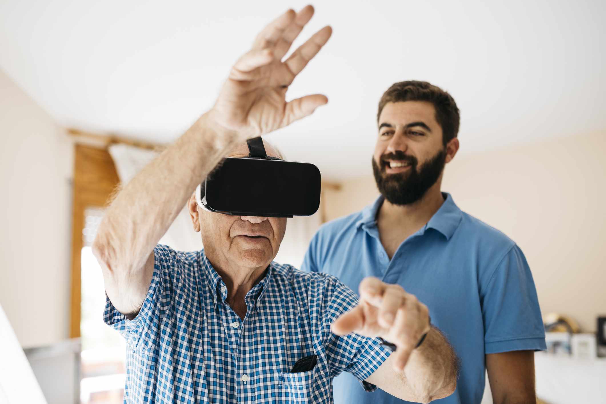 Starší muž doma pomocí brýlí virtuální reality, zatímco jeho dospělý vnuk ho sleduje