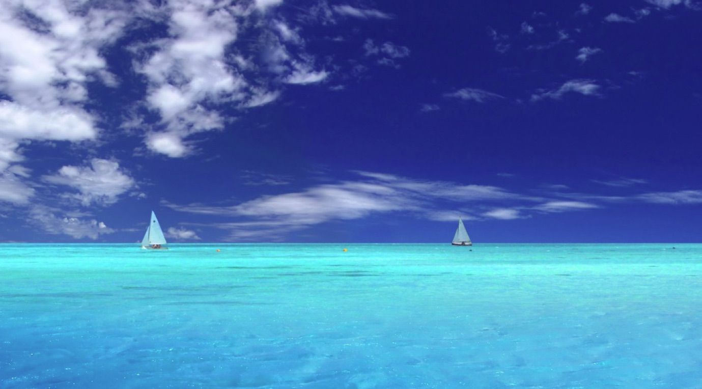 Zdarma oceánská tapeta se vzdálenými bílými plachetnicemi ve světle modrém oceánu pod modrou oblohou s rozptýlenými mraky