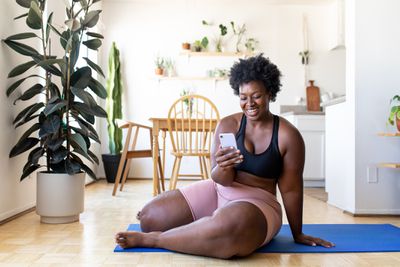 Žena sedí na podlaze v fitness zařízení se usmívá na její smartphone