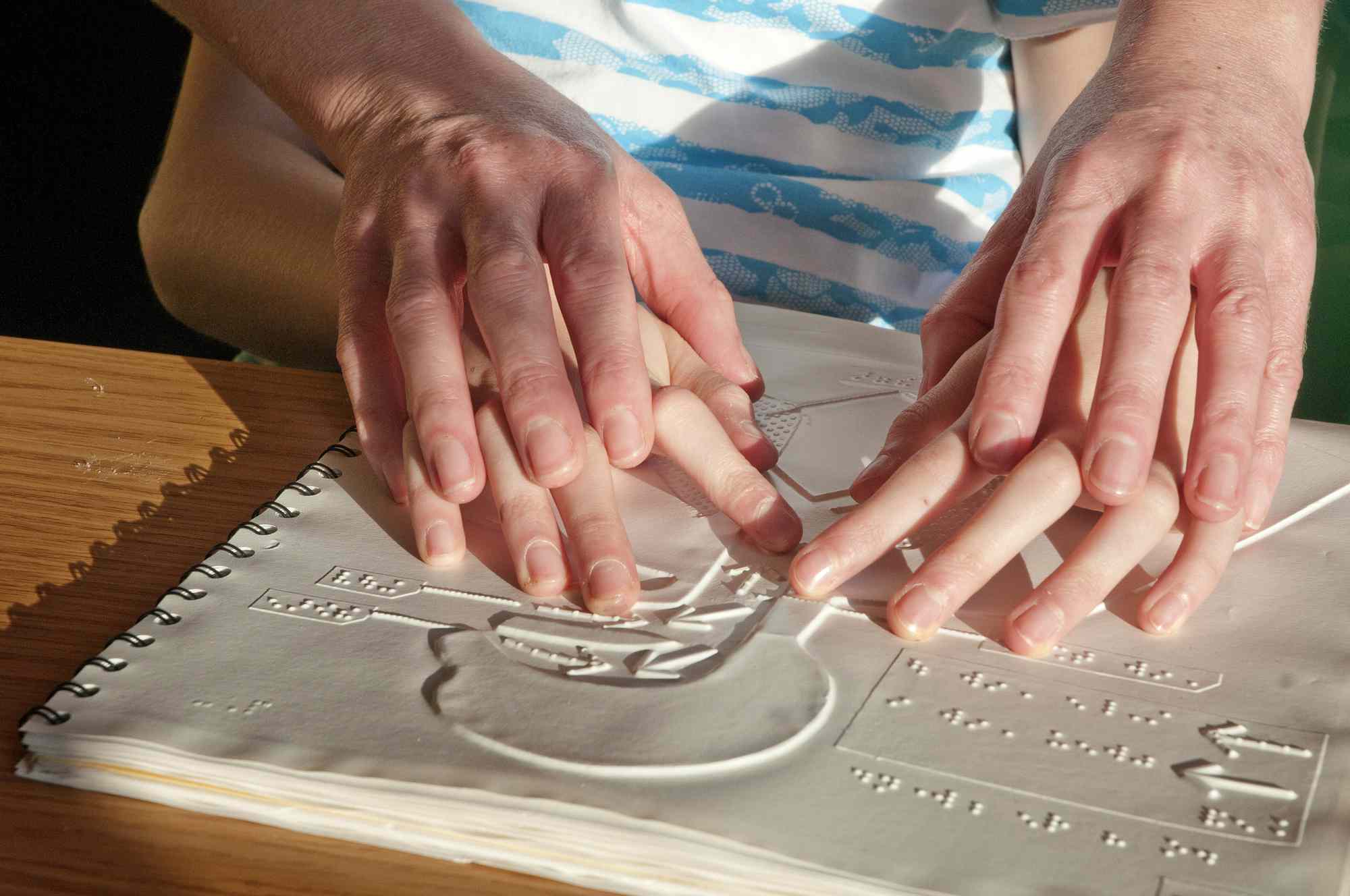 Detailní záběr na dospělé ruce, které vedou dětské ruce nad tiskem Brail v knize.