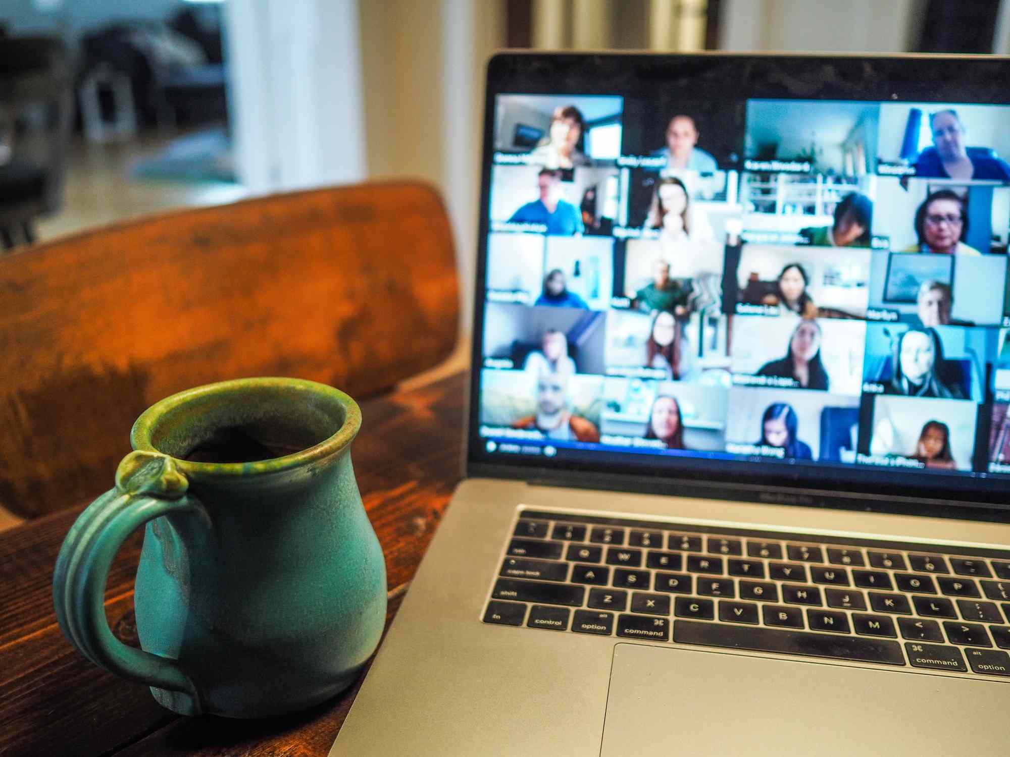 Otevřený notebook na kuchyňské desce zobrazující videohovor více osob s šálkem kávy poblíž. 